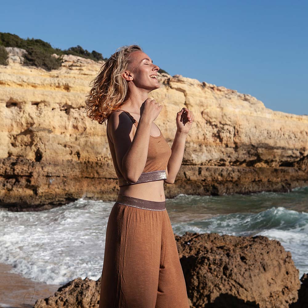 Lisa Rossmann seitlich vor Felsen am Strand, im Hintergrund die Wellen, wie sie sich den Wind durch die Haare wehen lässt und lächelnd ihr Gesicht mit geschlossenen Augen zur Sonne hebt.