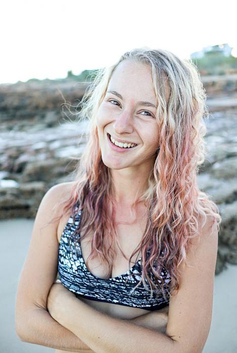 Nahaufnahme von Lisa Rossmann am Strand, im Hintergrund Felsen, wie sie mit unter der Brust verschränkten Armen in die Kamera lächelt.