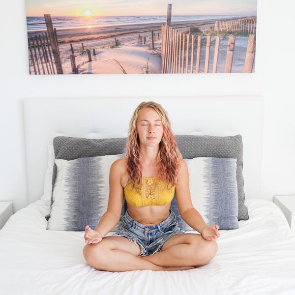 Lisa Rossmann sitzt in Jeanshorts und einem gelben Top mit geschlossenen Augen und gekreuzten Beinen auf einem Bett und meditiert. Im Hintergrund hängt ein Strand-Bild mit Dünen und Sonnenuntergang.